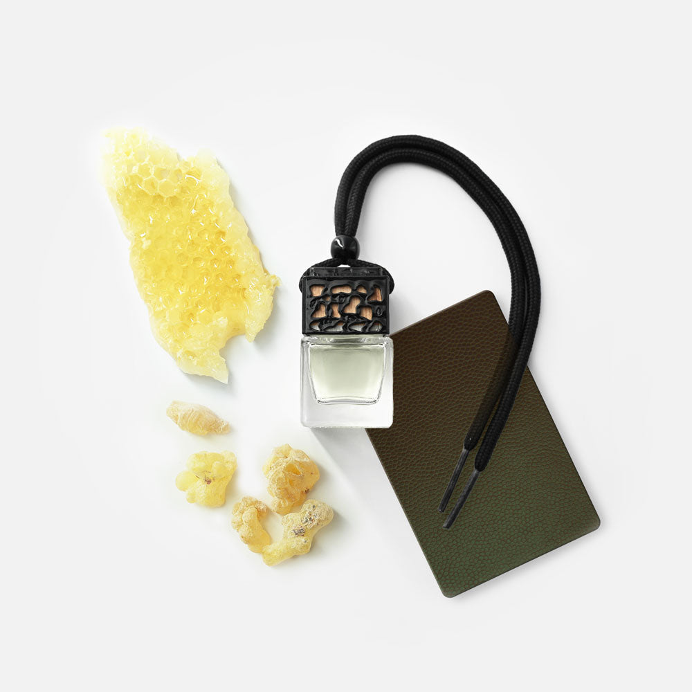 Ein kleines Glasfläschchen mit schwarzem Verschluss, daran eine schwarze Kordel, daneben ein Stück Bienenwabe, Harzstücke und eine rechteckige, dunkelgrüne Karte auf weißem Hintergrund.